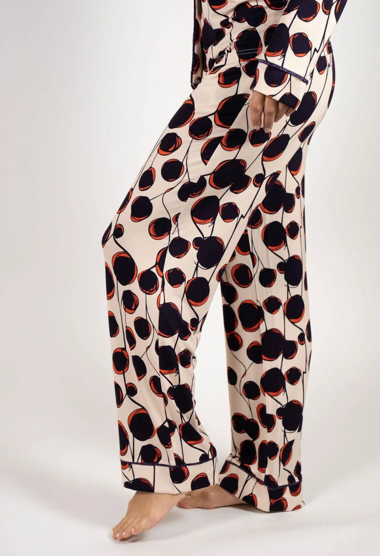 A person wearing a matching long-sleeve pajama top and wide-leg pants set with an abstract floral pattern in black, dark navy, and orange-red on a warm cream background, photographed against a light gray studio backdrop.