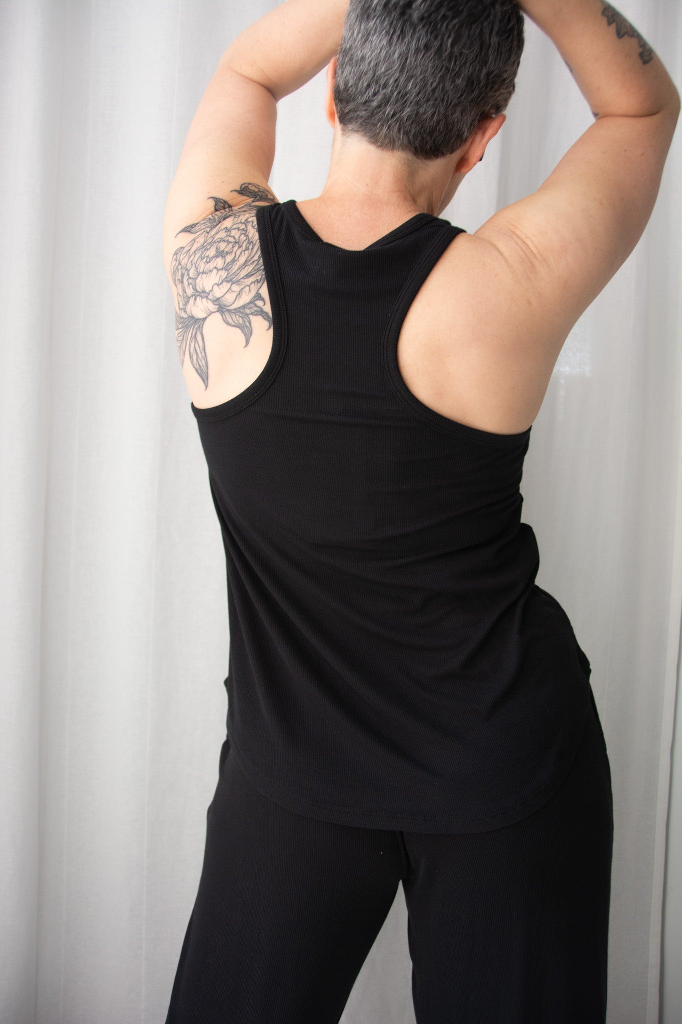 Person wearing a black tank top and pants with tattoos on arms against a white curtain background