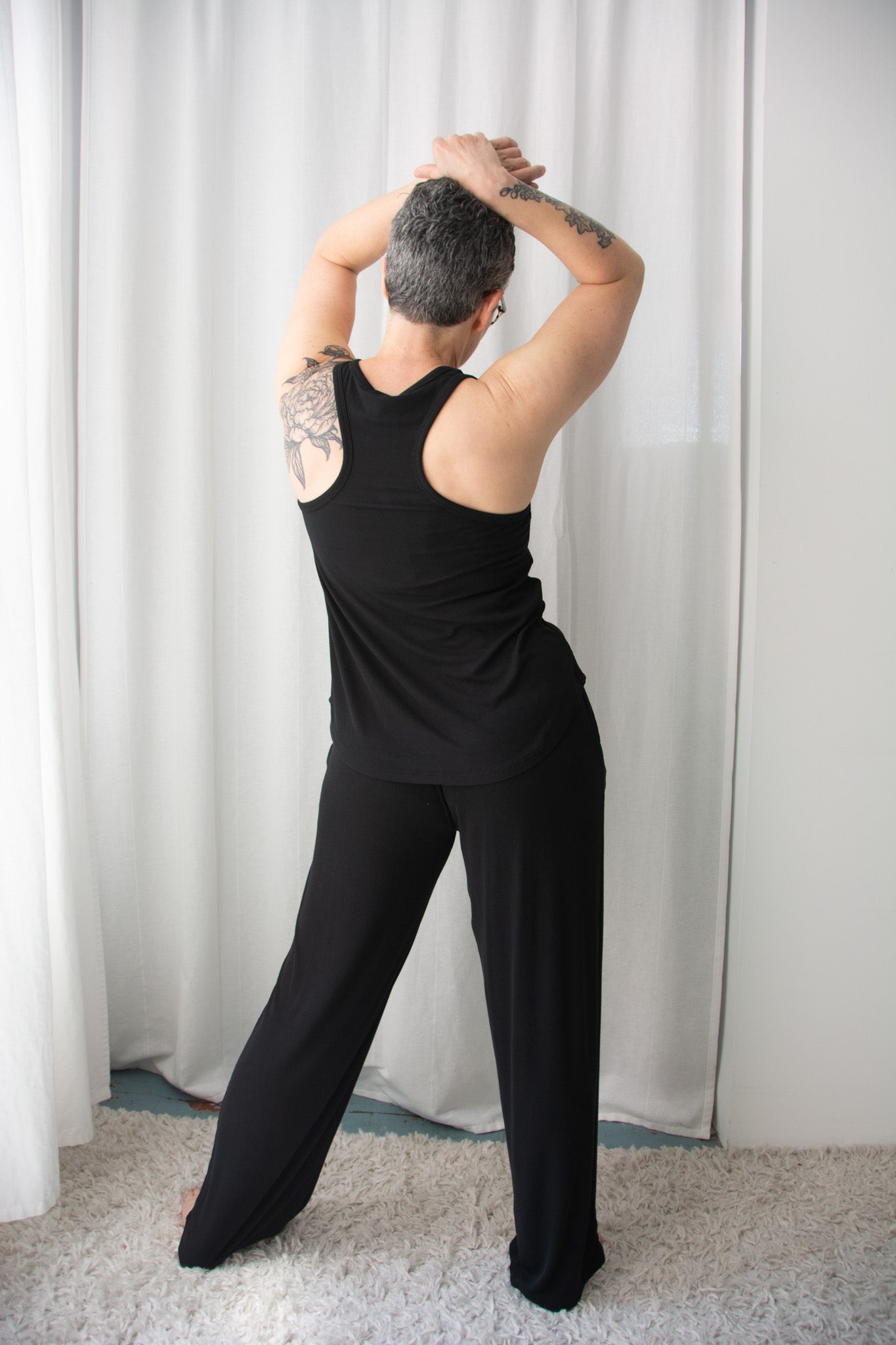 Person wearing a black tank top and pants stretching against a white curtain.