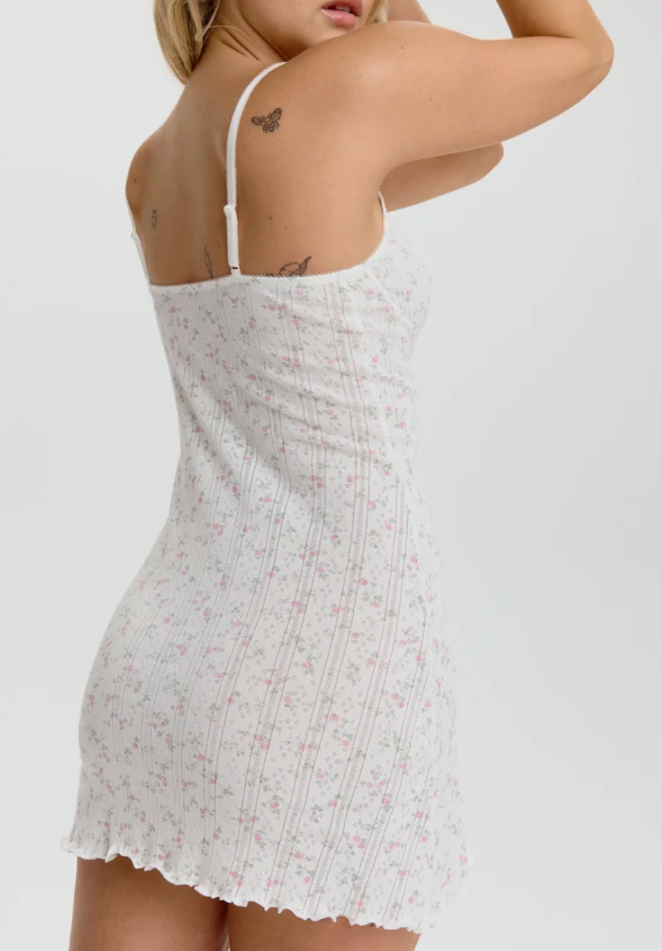 A woman wearing a white ribbed mini dress with a delicate pink and green floral print, shown from the back against a plain white studio background.
