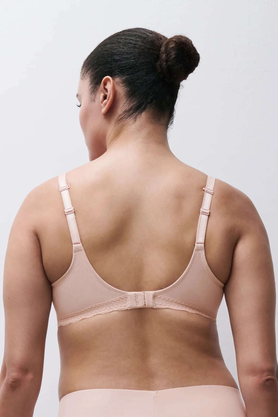 Rear view of a woman wearing a light beige wireless bra and matching underwear against a plain light gray background.