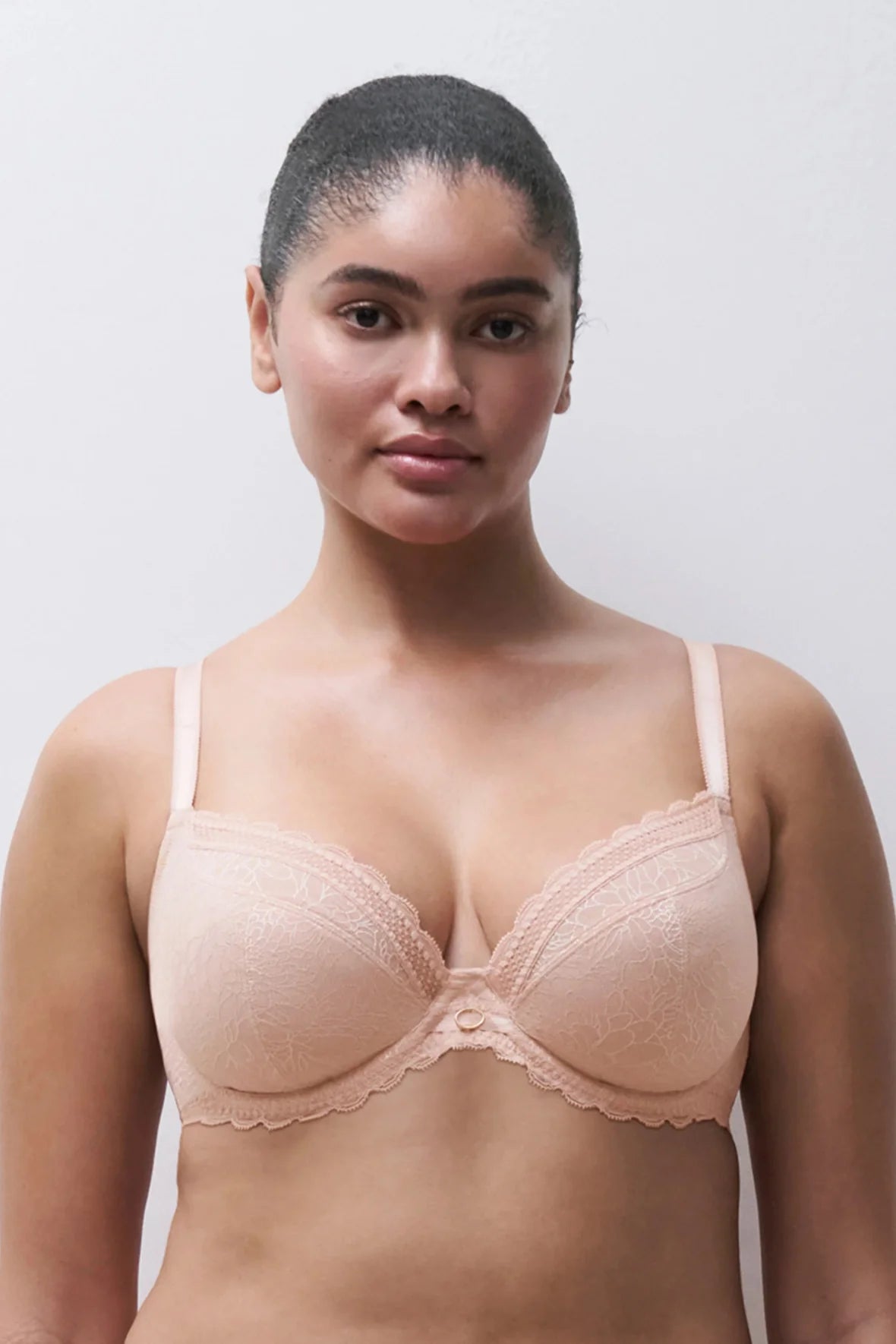 A woman modeling a light peach-beige lace underwire bra against a plain light gray background.