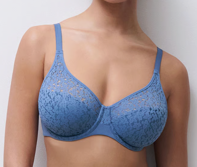 A woman wearing a blue lace underwire bra against a plain light gray wall.