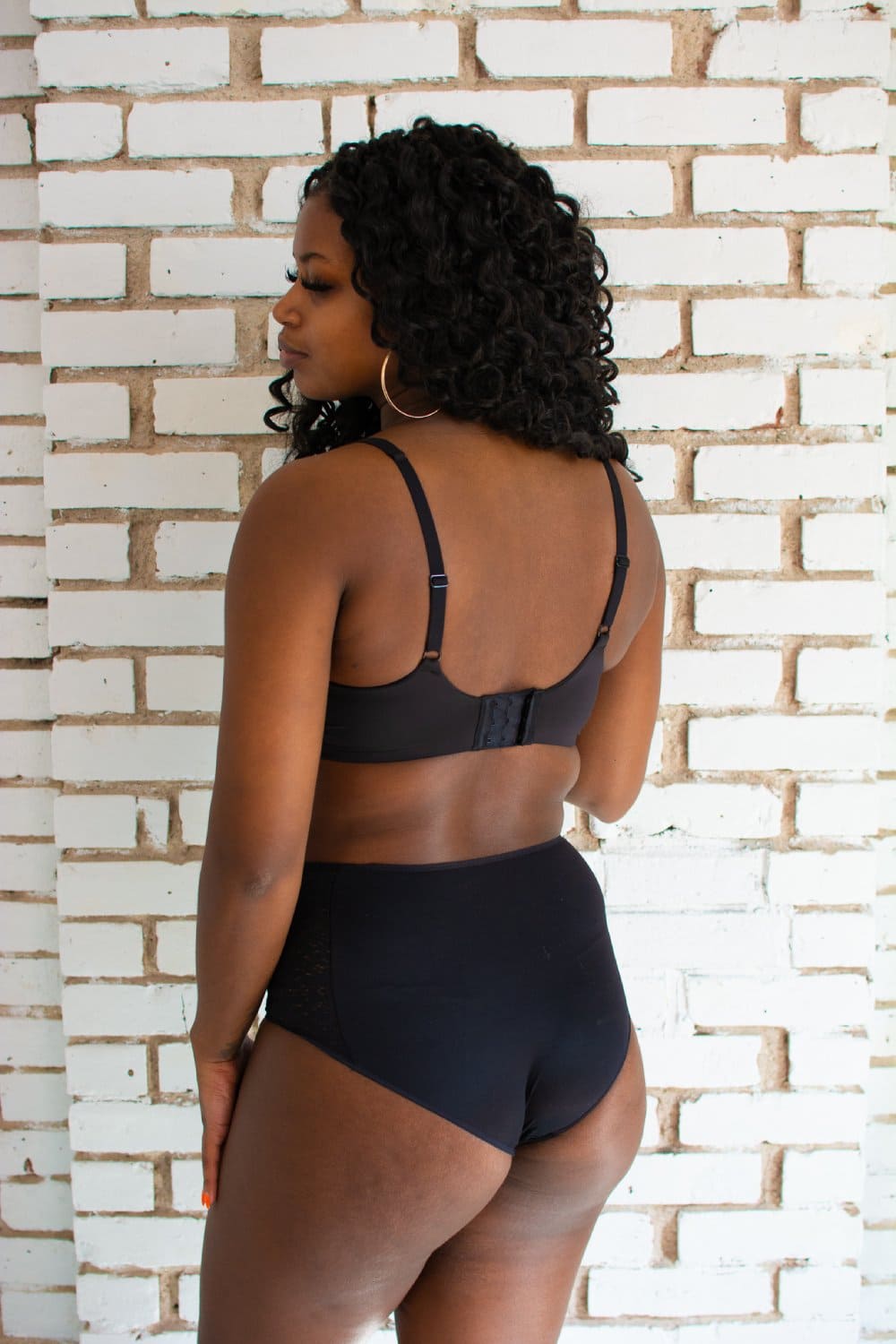 Chantelle norah high waist brief in black, back view on a model, standing at an angle, brick wall background.