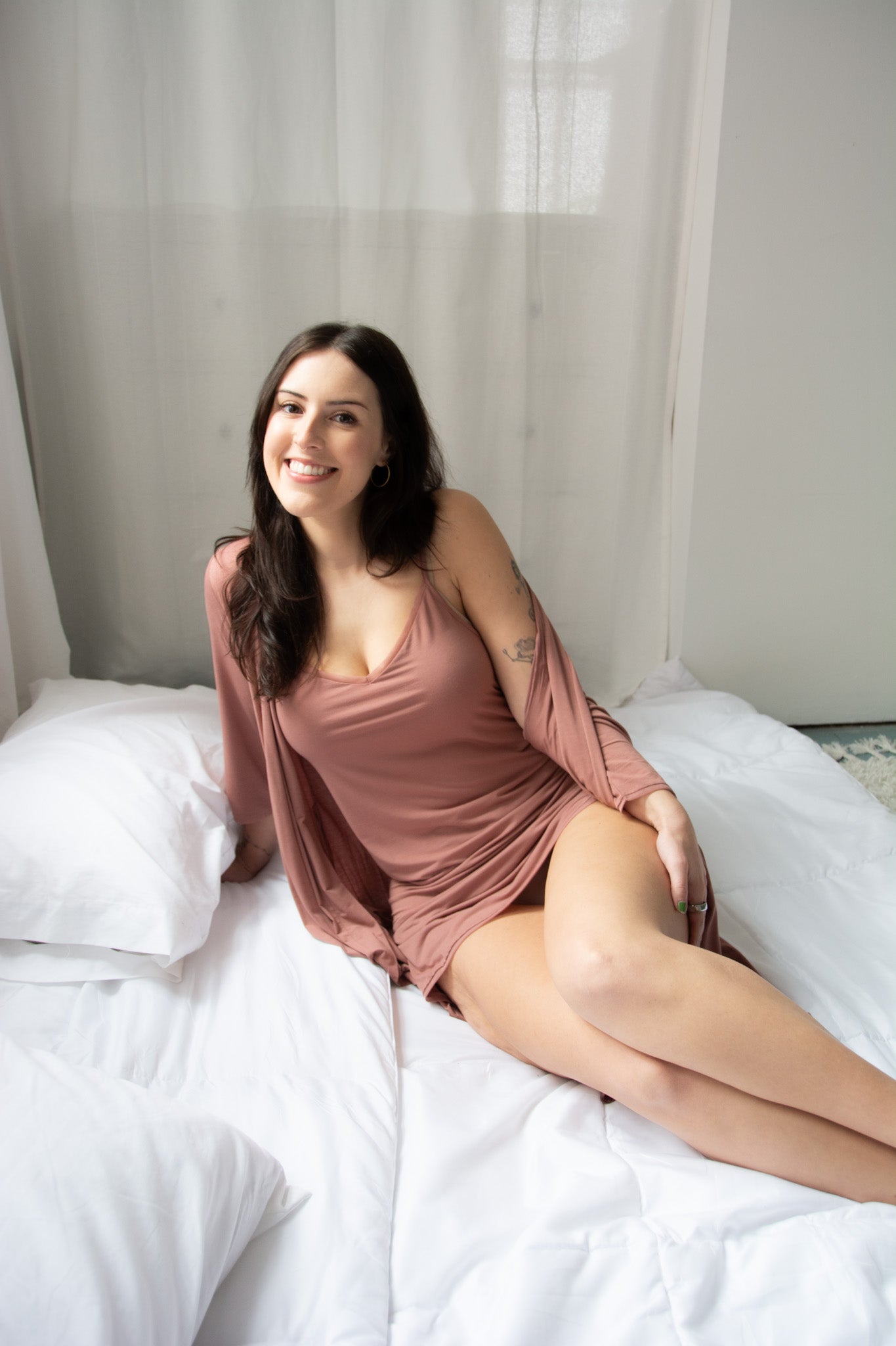 Model wearing dusty rose chemise and robe on a white bed.