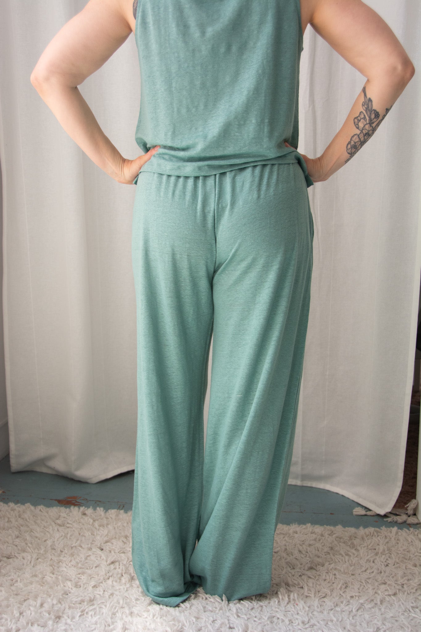 Person wearing a teal sleeveless top and wide-leg pants against a white curtain background