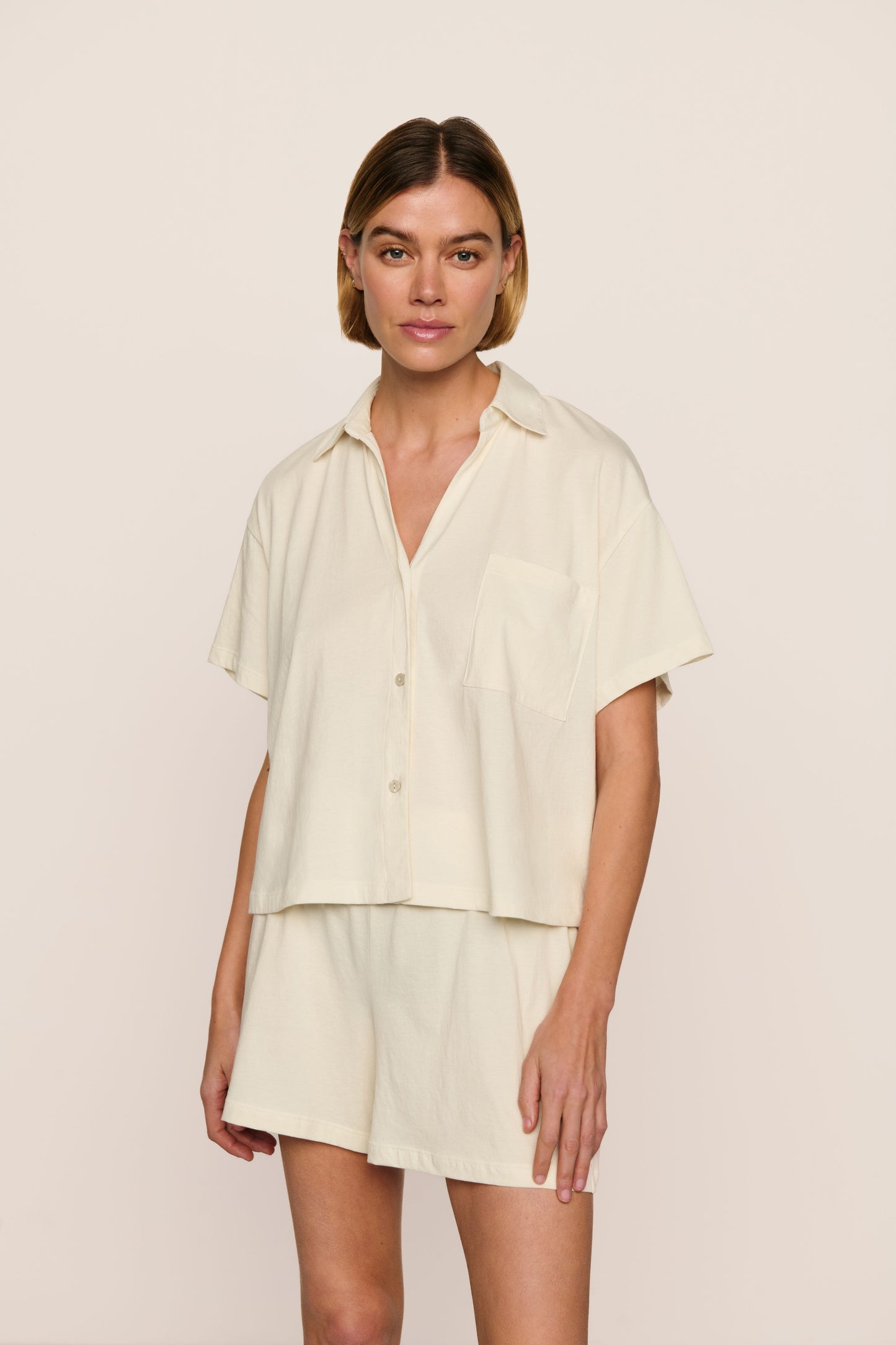 A woman modeling a matching two-piece lounge set consisting of a short-sleeve button-front shirt and shorts in a light cream color, set against a plain beige studio background.
