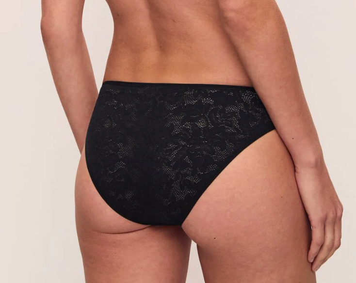 Rear view of a model wearing black lace high-waisted briefs against a plain light beige studio background.