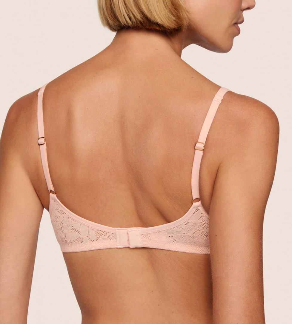 A woman shown from the back wearing a light pink lace bra with thin adjustable straps and a center tie, against a plain light beige studio background.