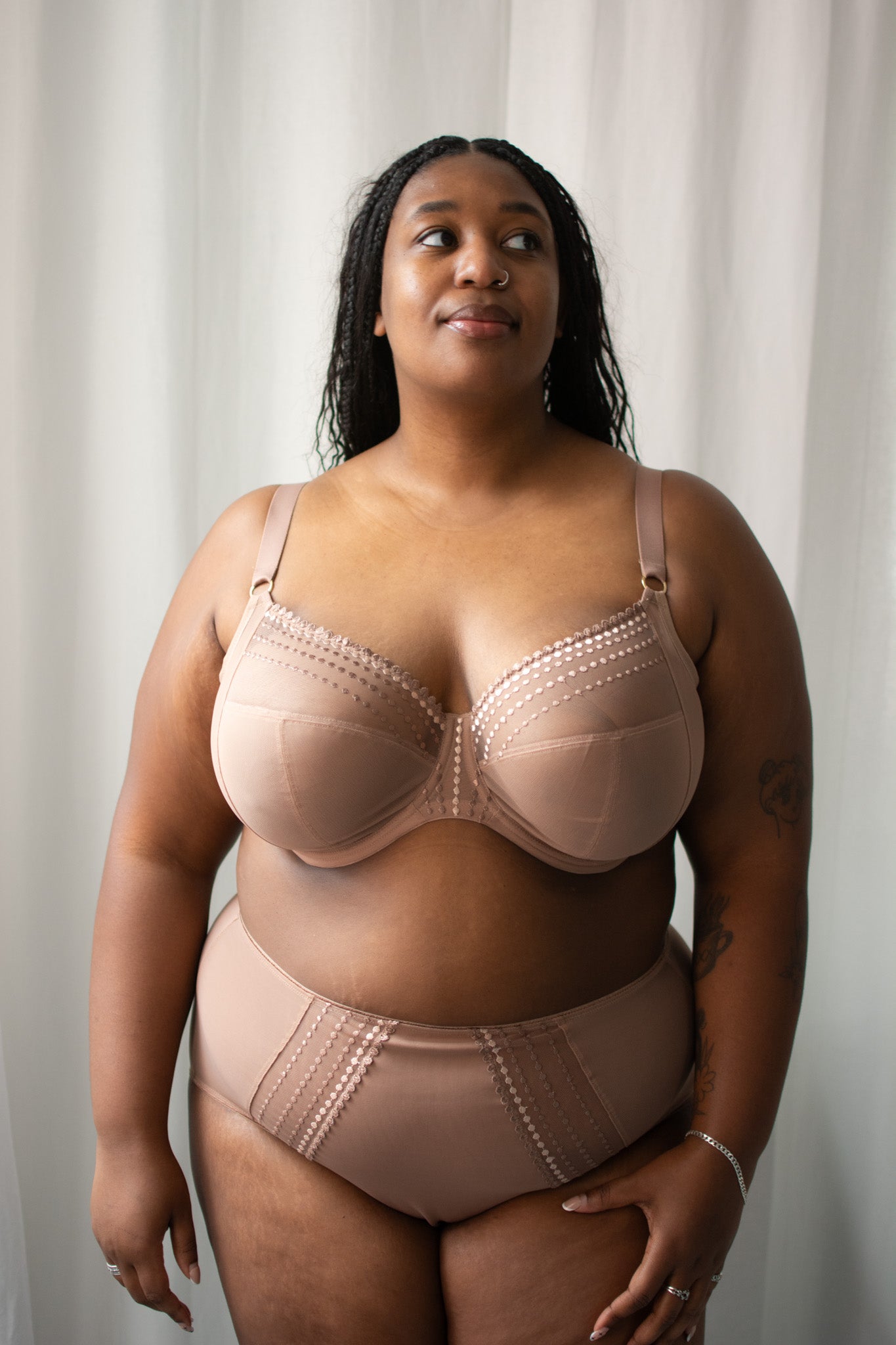 Woman wearing a beige bra and panties set against a white curtain background