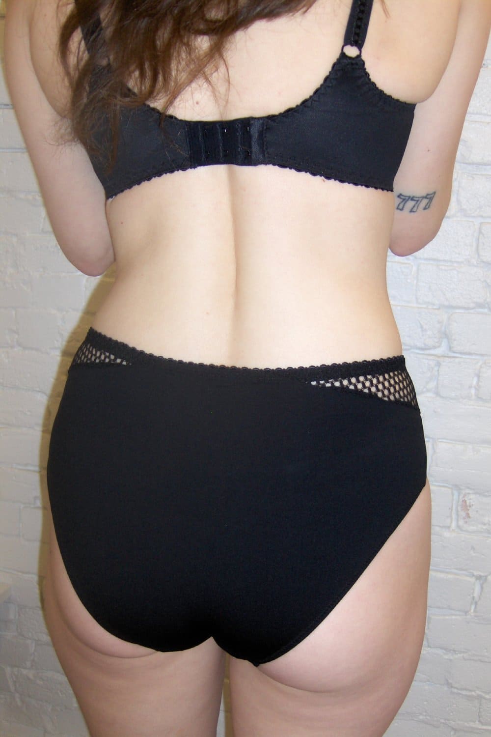 Person wearing a black bra and panties set against a light gray brick wall.