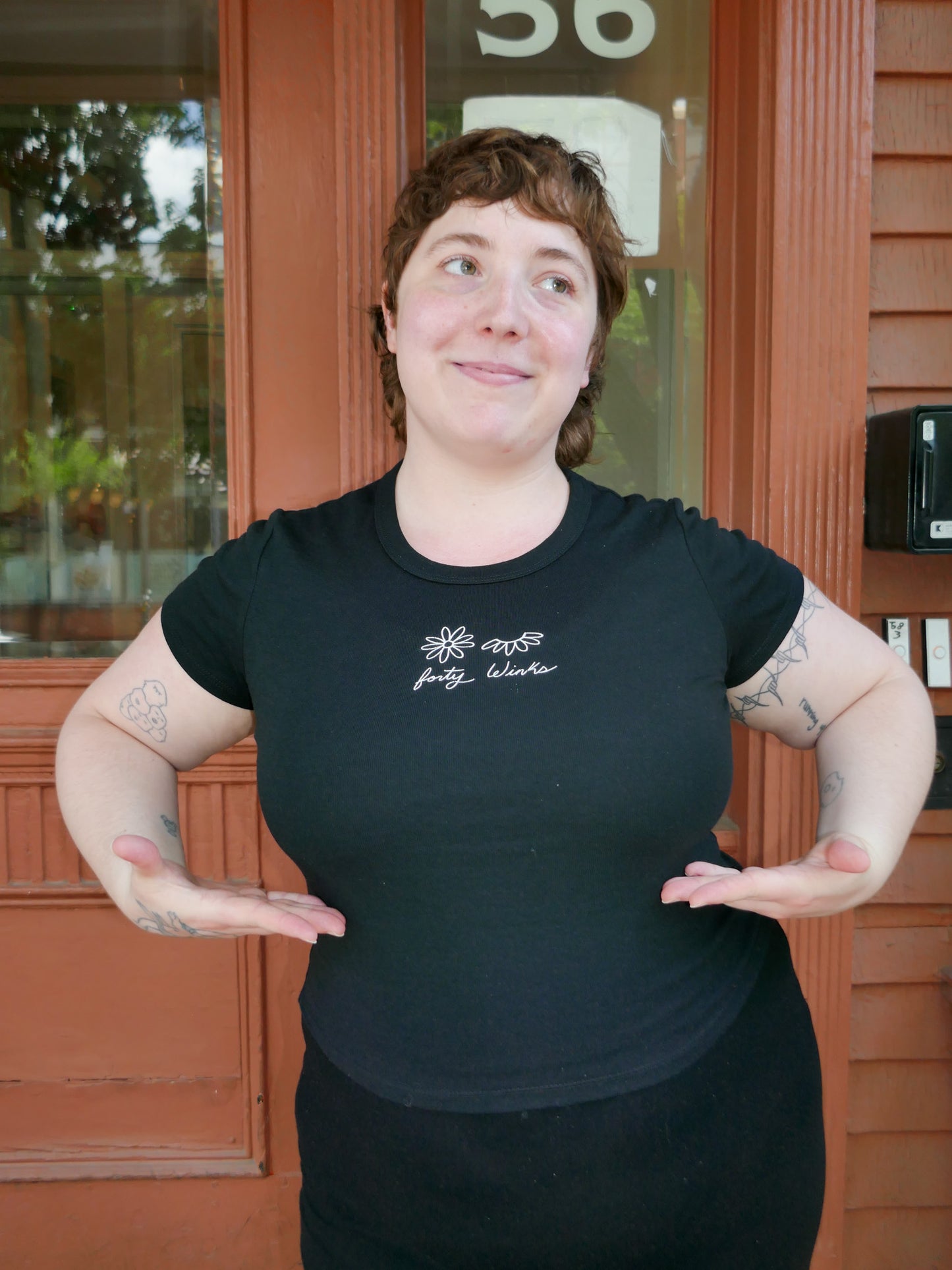 Person wearing a black t-shirt with a logo and text, standing in front of a door.