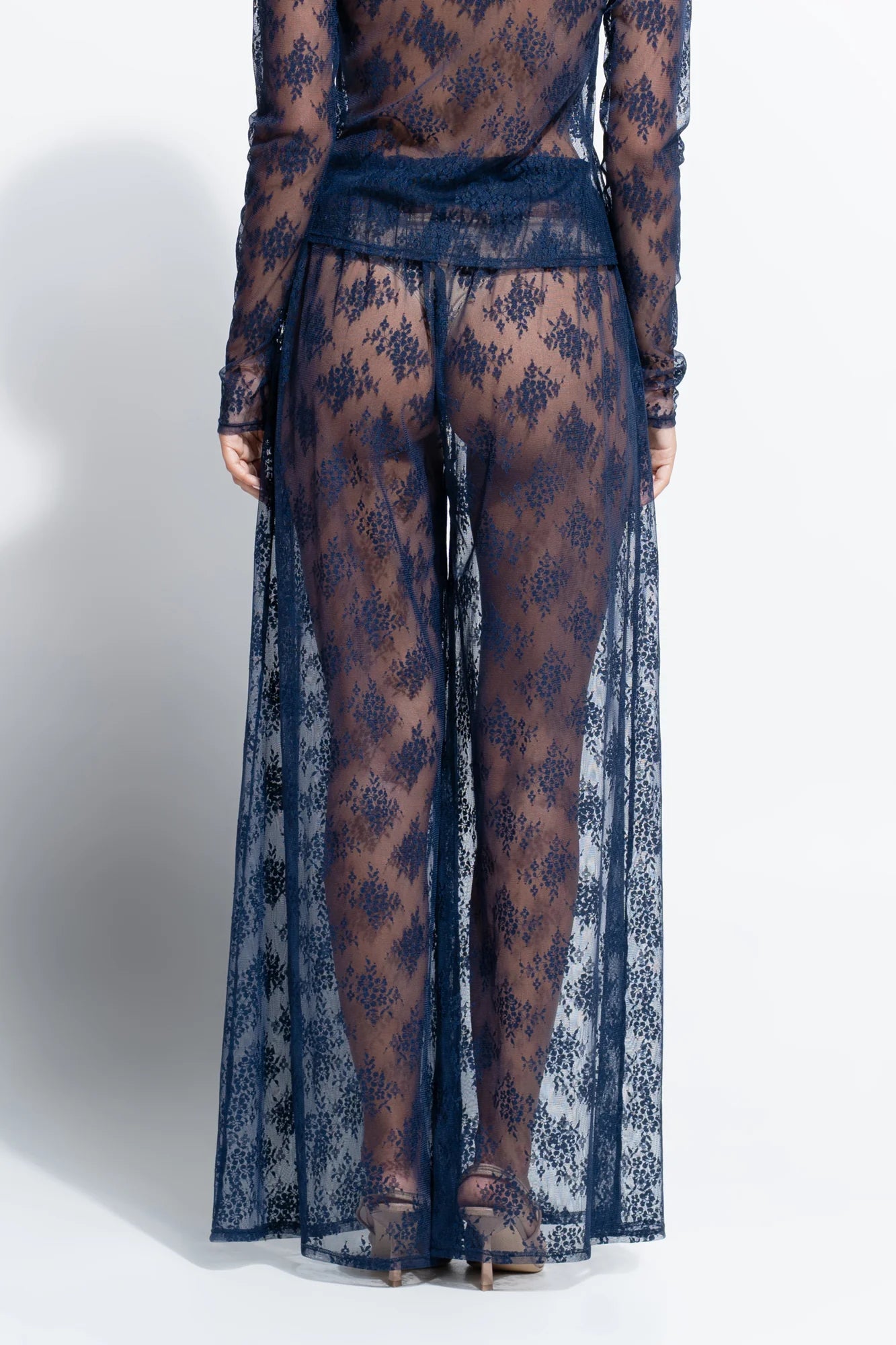 A model shown from the back wearing a sheer navy blue lace long-sleeve top and matching wide-leg pants with a long draped overlay, photographed against a light gray studio background.
