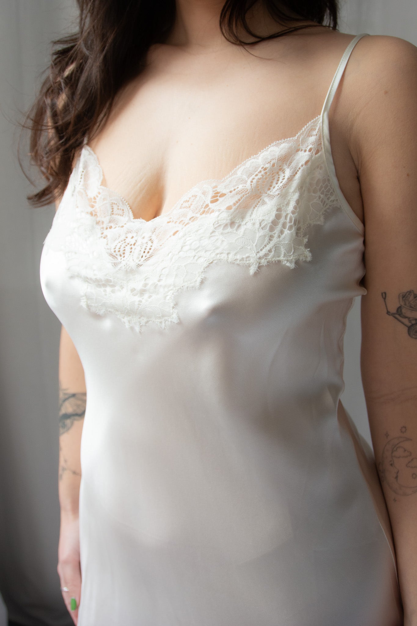 A woman wearing a white silk chemise with lace detailing at the neckline and mid-thigh length. She is holding a bouquet of flowers and standing against a white background.