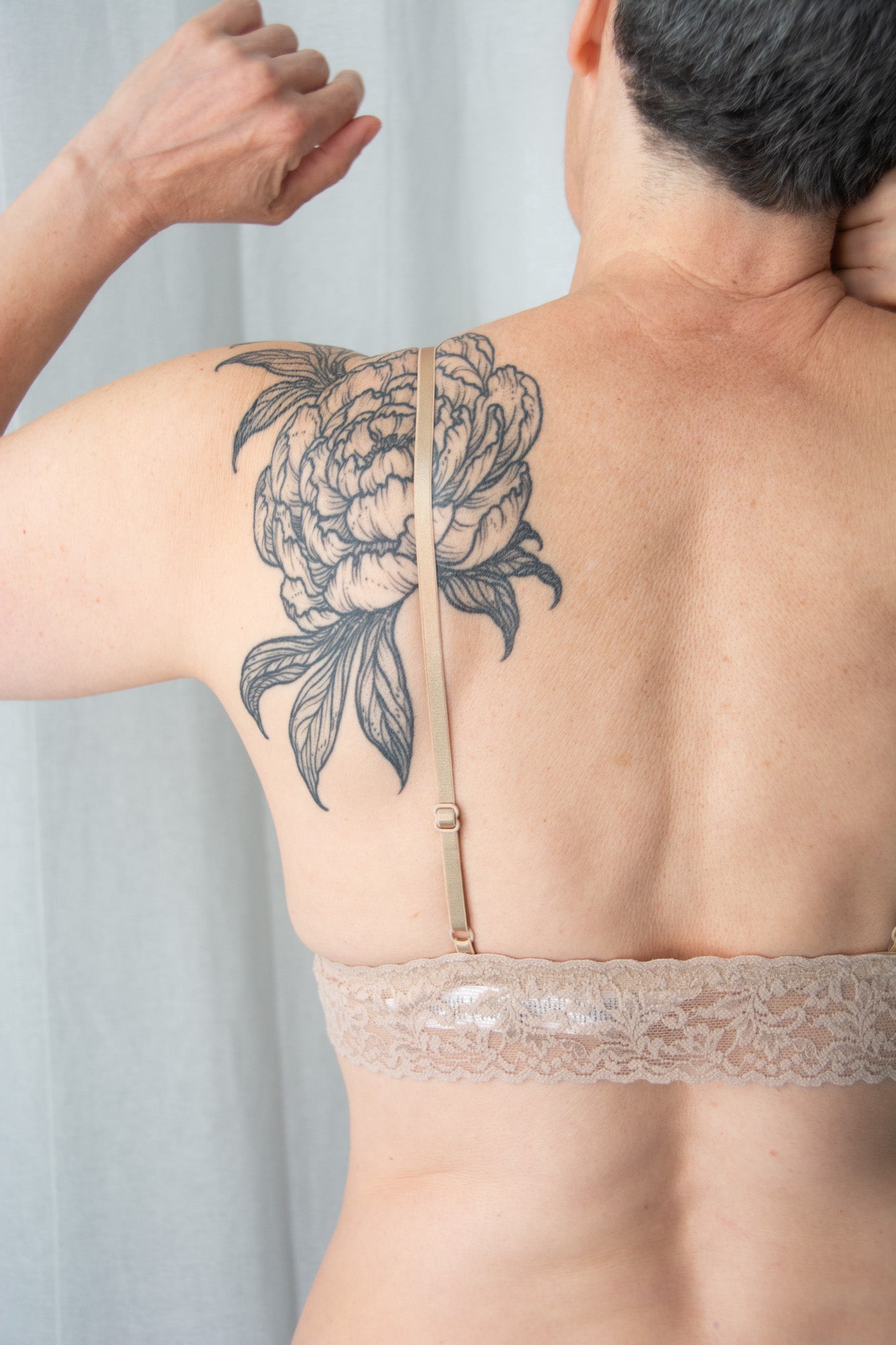 Person wearing a beige lace bra with a floral tattoo on the back against a light gray curtain.