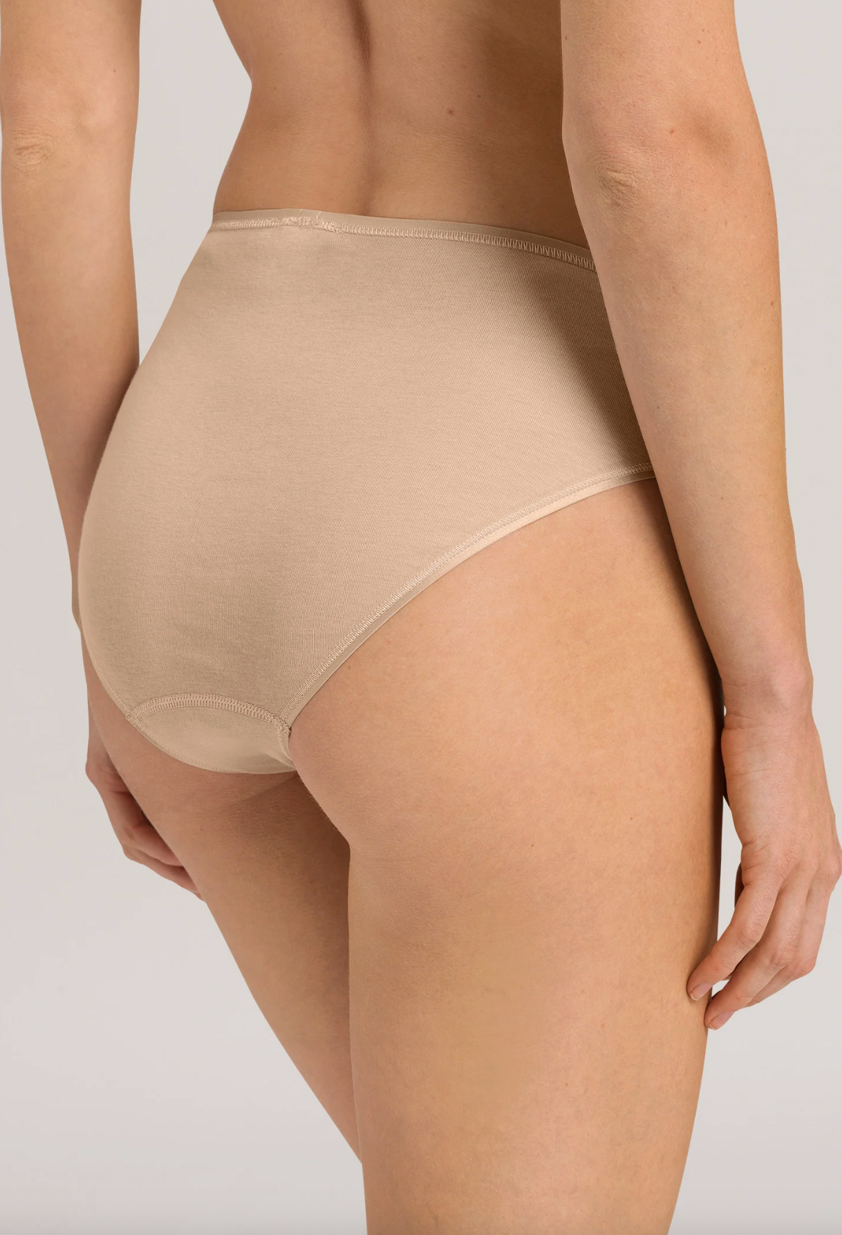 A close-up back view of a model wearing high-waisted beige briefs against a light gray studio background.