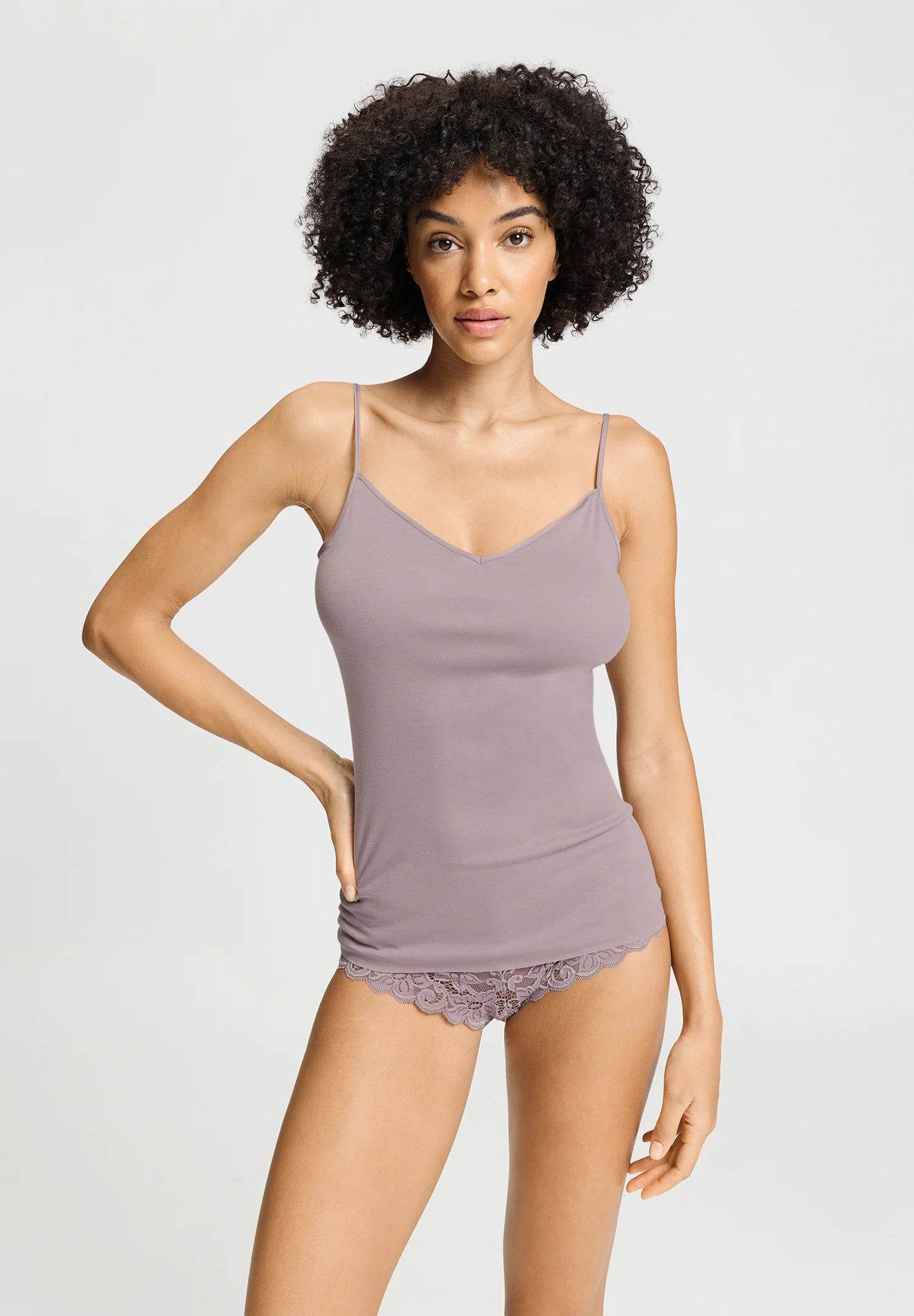 A model with curly dark hair wearing a light dusty purple camisole and matching lace-trimmed underwear against a plain light gray studio background.