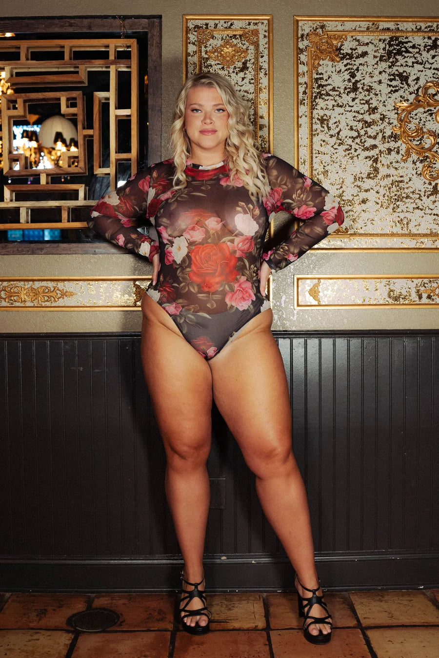 woman wearing mesh floral long sleeve bodysuit with black heels against a wall with gold detail.