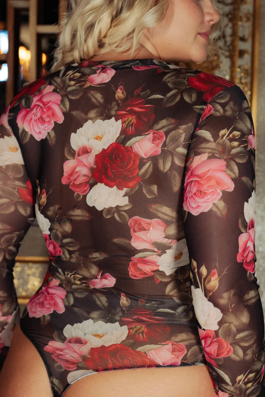 back of woman wearing mesh floral long sleeve bodysuit against a wall with gold detail.