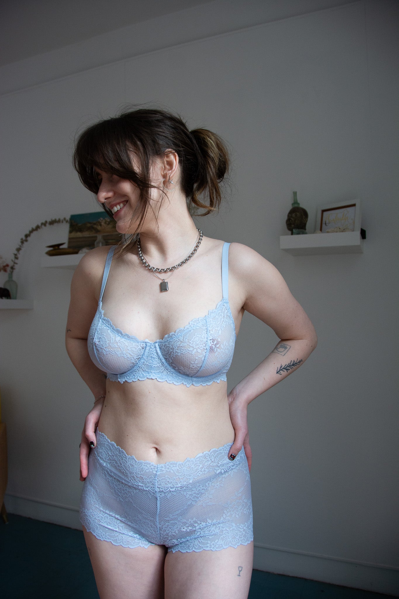 person wearing blue lace bra and underwear in a white room with shelves.