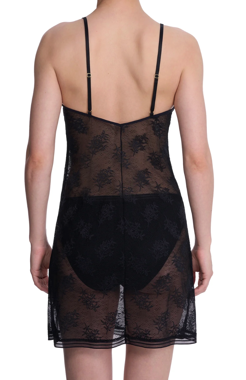 A woman shown from the back wearing a black sheer lace slip dress with thin straps and a floral lace pattern, photographed against a plain white studio background.