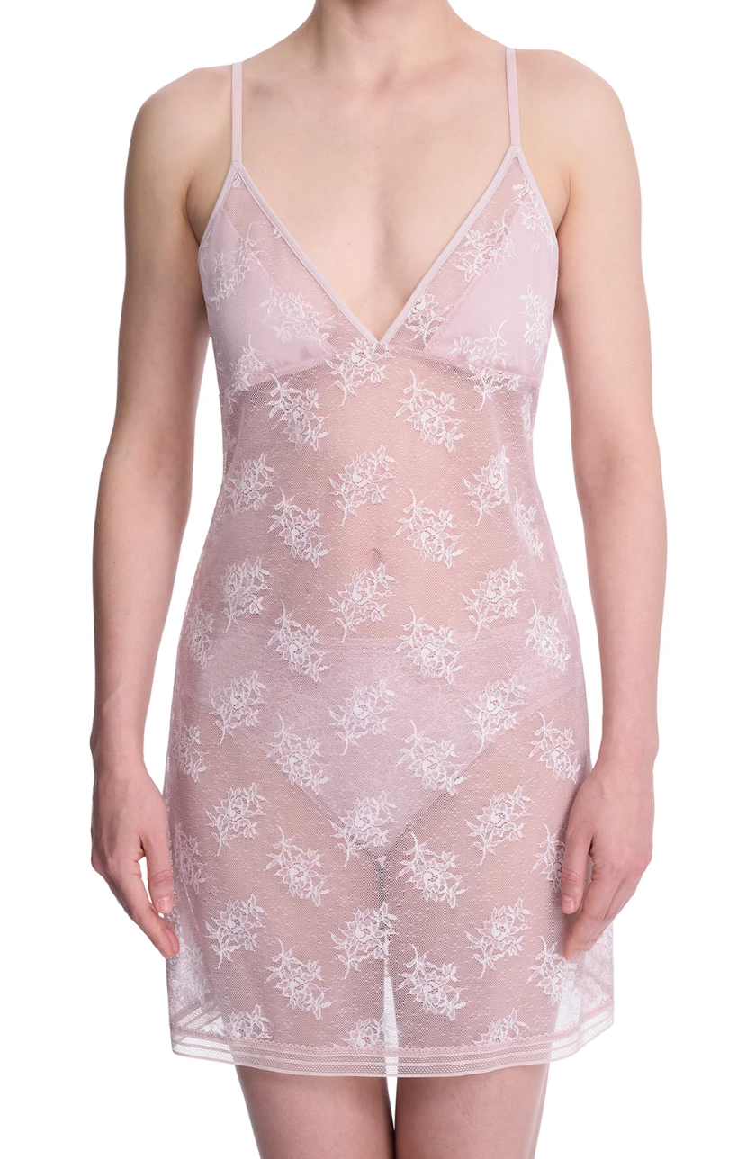 A woman wearing a sheer pale pink lace slip dress with white floral embroidery, photographed against a plain white studio background.
