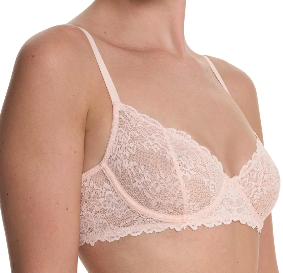 A close-up of a model wearing a pale pink lace bra with thin straps and a scalloped floral lace cup design, shown against a plain white background.