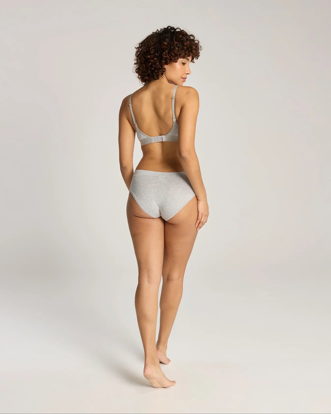 A woman with curly brown hair is shown from behind wearing a light gray two-piece lingerie set, consisting of a bralette and high-waisted briefs, against a plain light gray studio background.