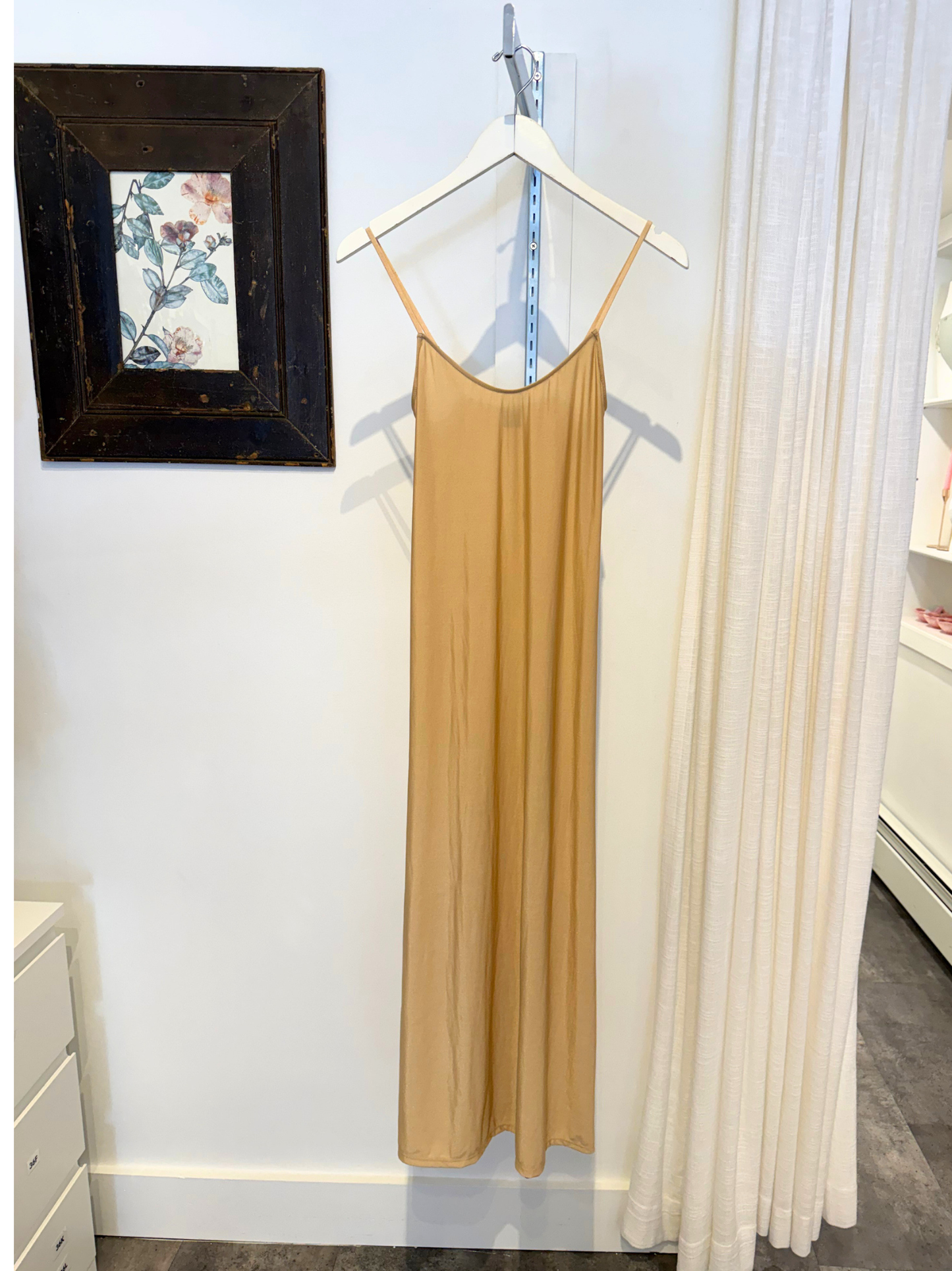 Beige slip dress hanging on a hanger against a white wall with a framed picture and curtains.
