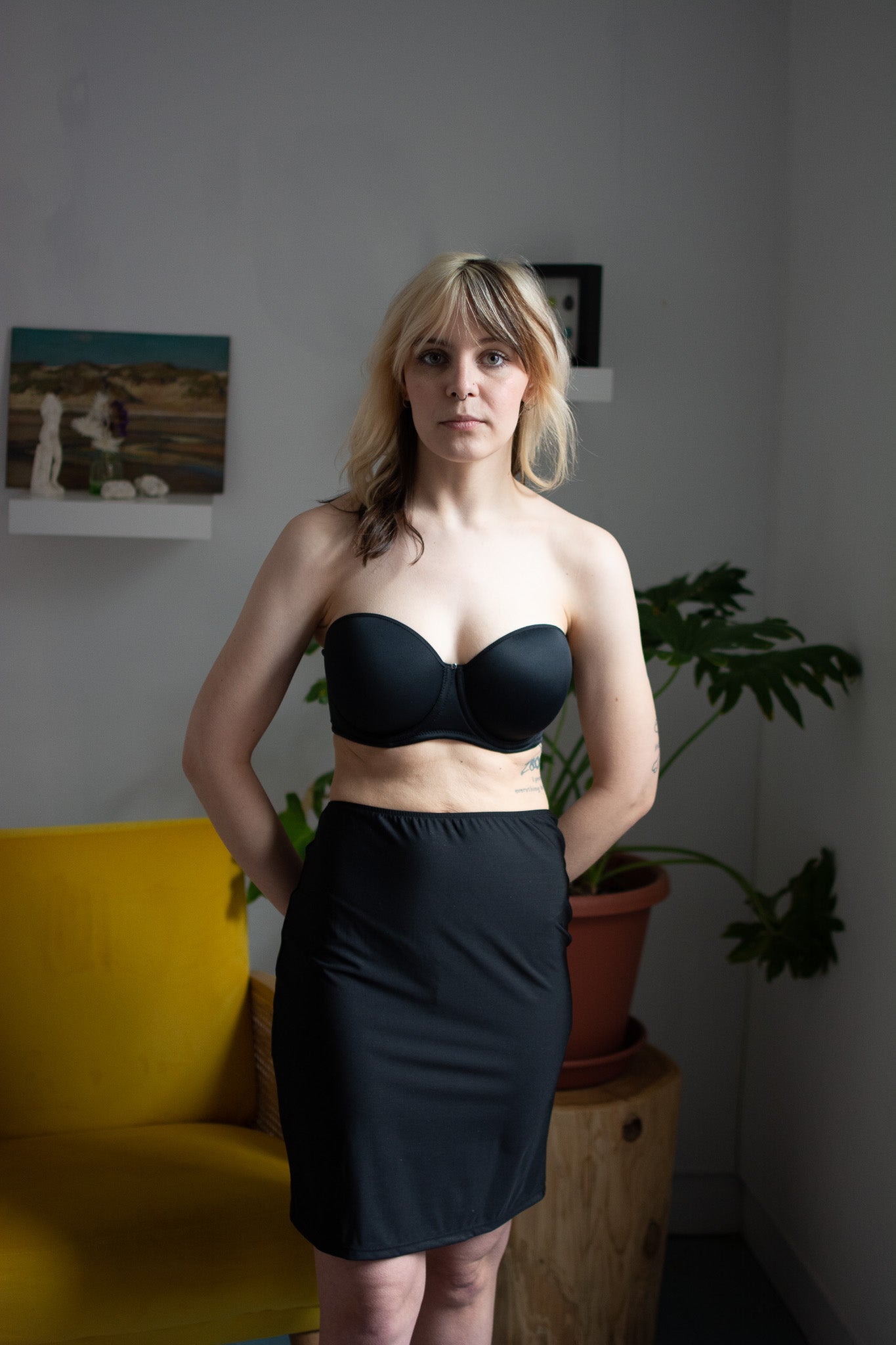 Woman wearing a black strapless top and skirt in a room with a yellow chair and potted plant.