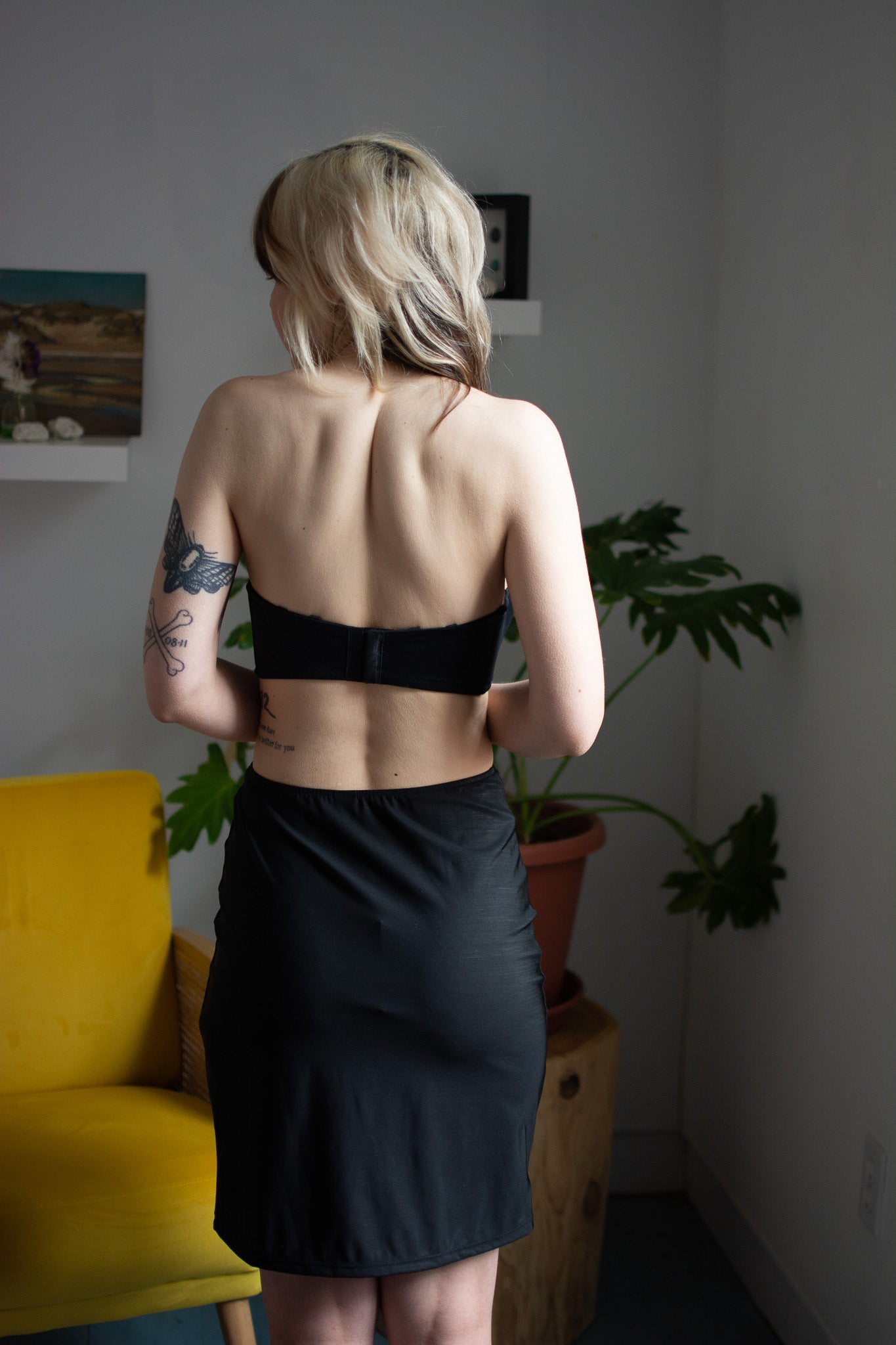 Person wearing a black strapless top and black skirt in a room with a yellow couch and plants.