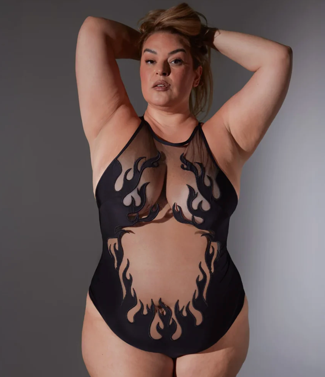 A plus-size model wearing a black sheer mesh bodysuit with a flame print, posing against a neutral gray studio background.