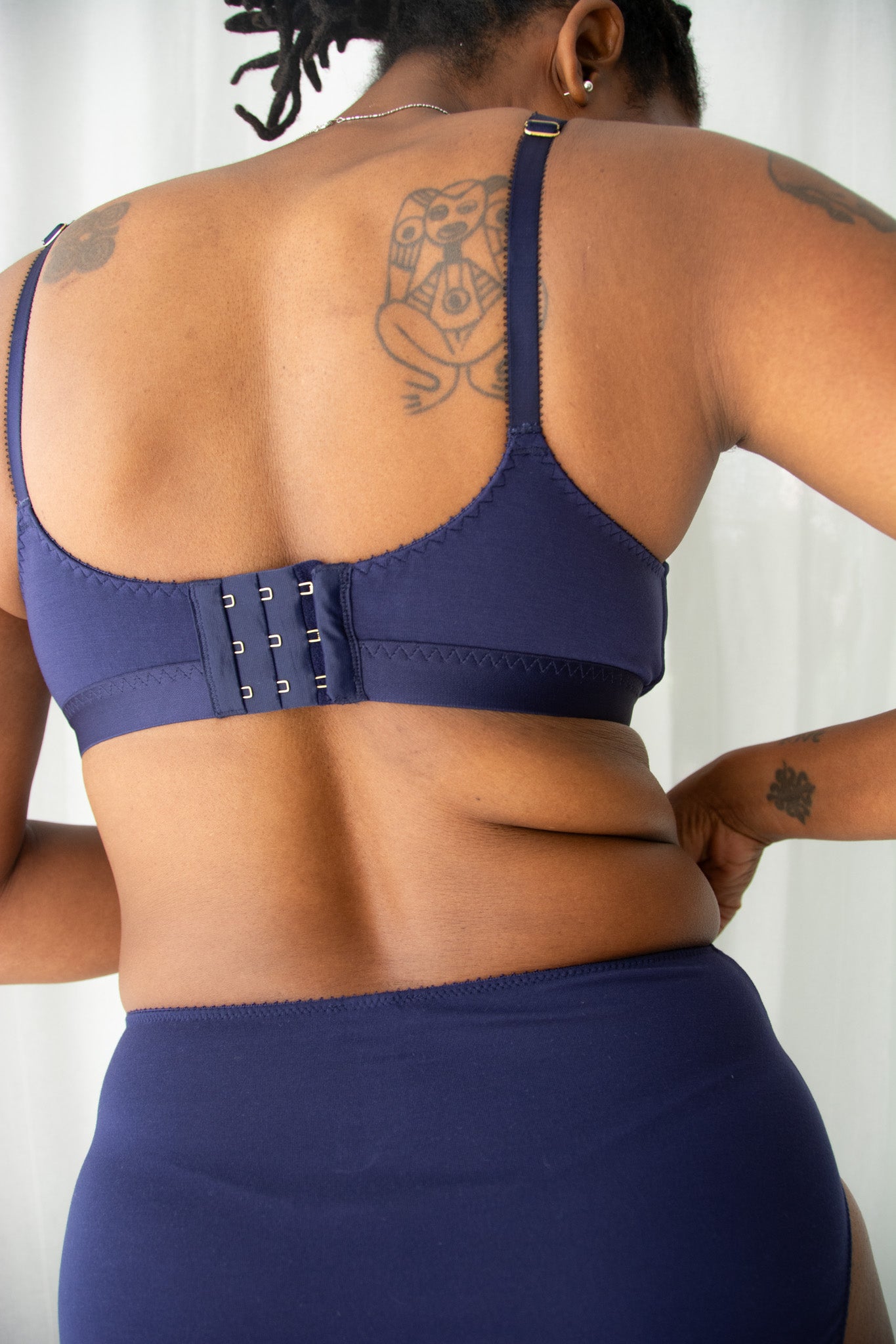 Back of a woman wearing a navy bra and underwear against a white curtain background