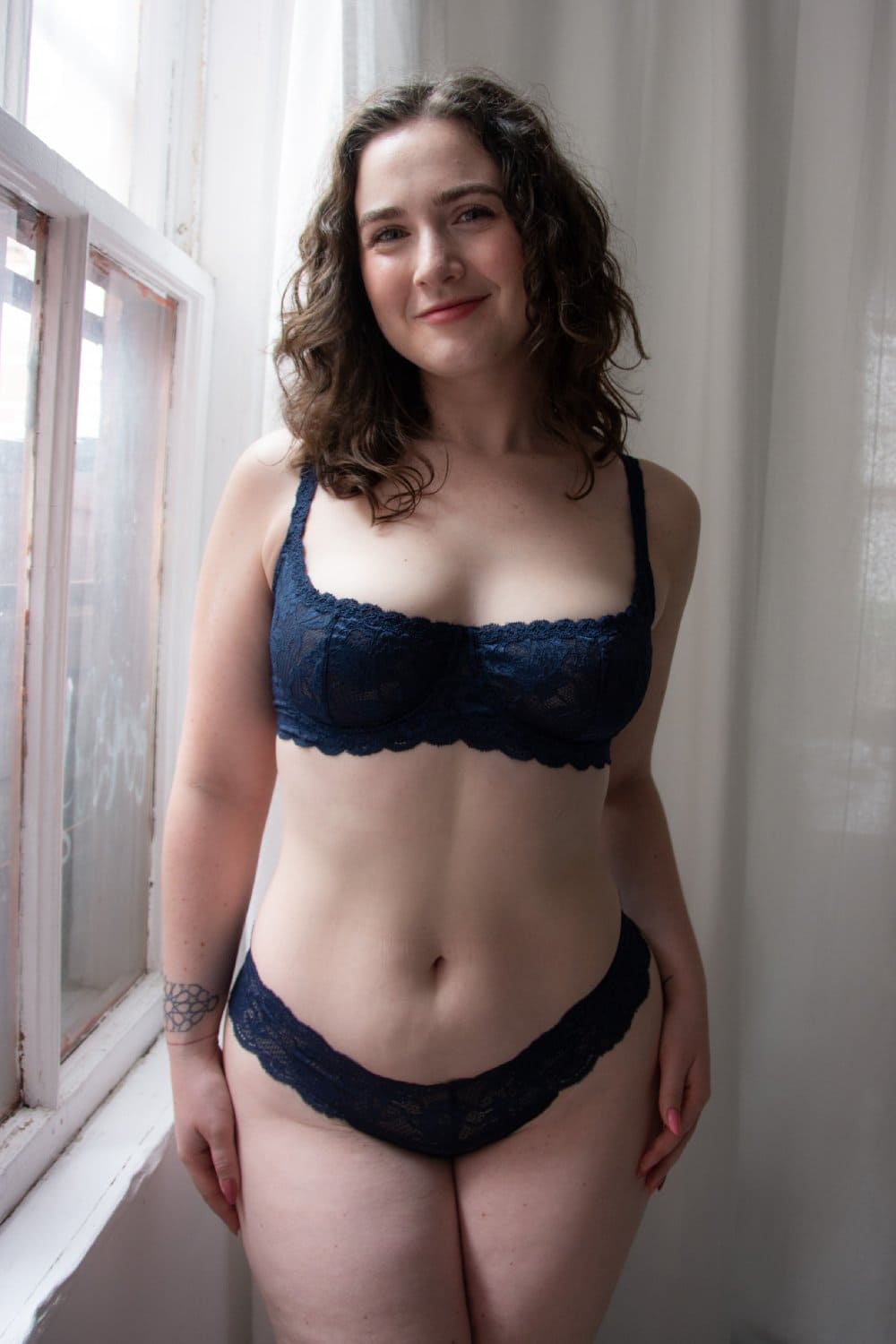 Woman wearing a navy blue lace bra and panties set in a softly lit room.